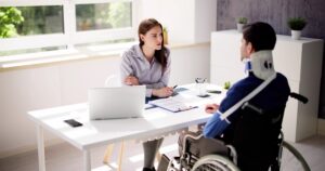 A lawyer meeting with an injured client in a wheelchair, with an arm sling, and a neck brace to discuss the timeline for their 18-wheeler lawsuit.