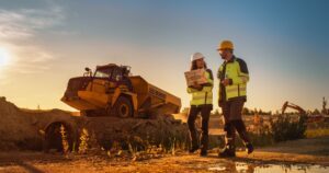 A man and woman wearing high-visibility vests and hard hats walk in front of a large yellow construction vehicle while discussing how liability works in construction truck accidents.